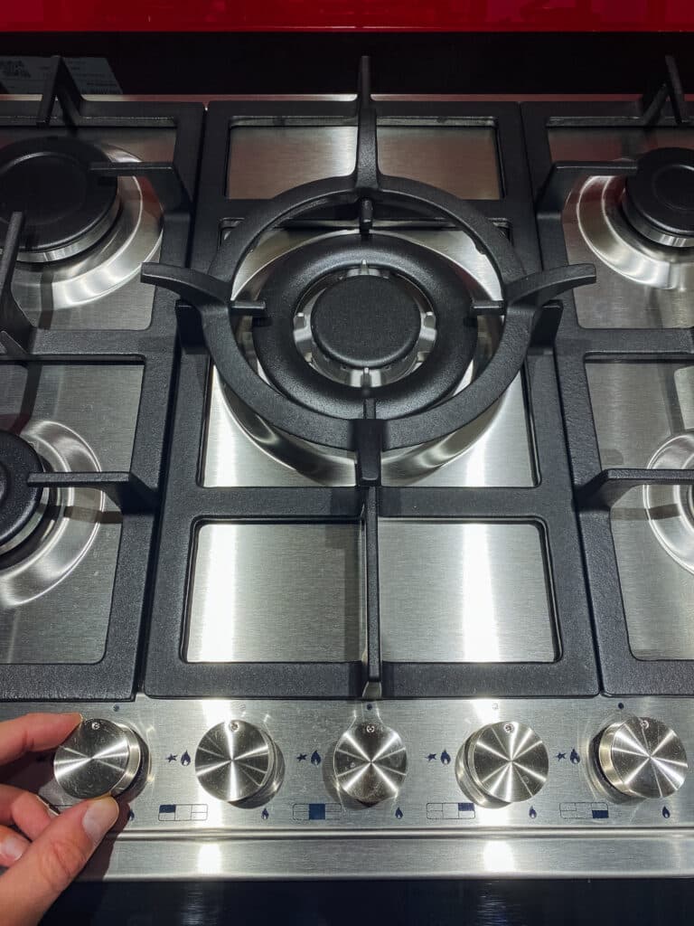 Four à Gaz Inox 5 Brûleurs : Réglage des Commandes Main ajustant le bouton d'un fourneau à gaz inox à cinq brûleurs. Le grand brûleur central est visible au-dessus des commandes.